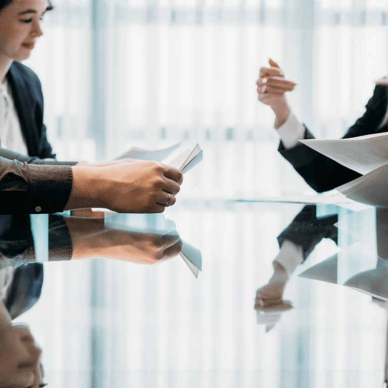 Two professionals seated at a glass table reviewing and exchanging documents during a business meeting