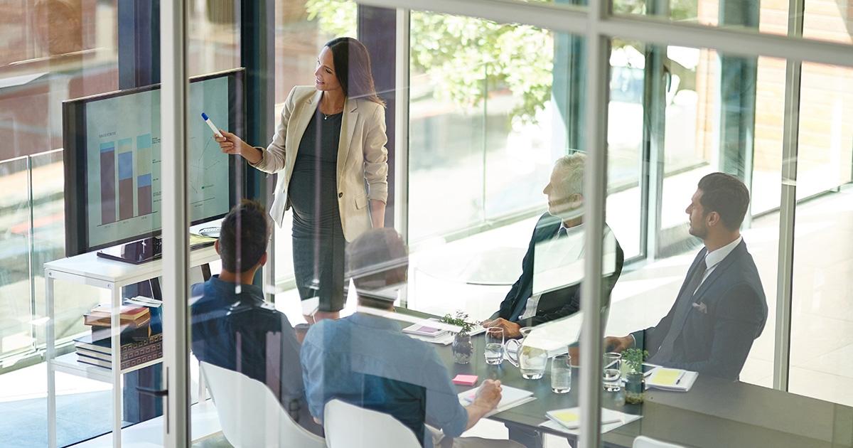 people in a board room listening to someone present at the front pointing to a screen
