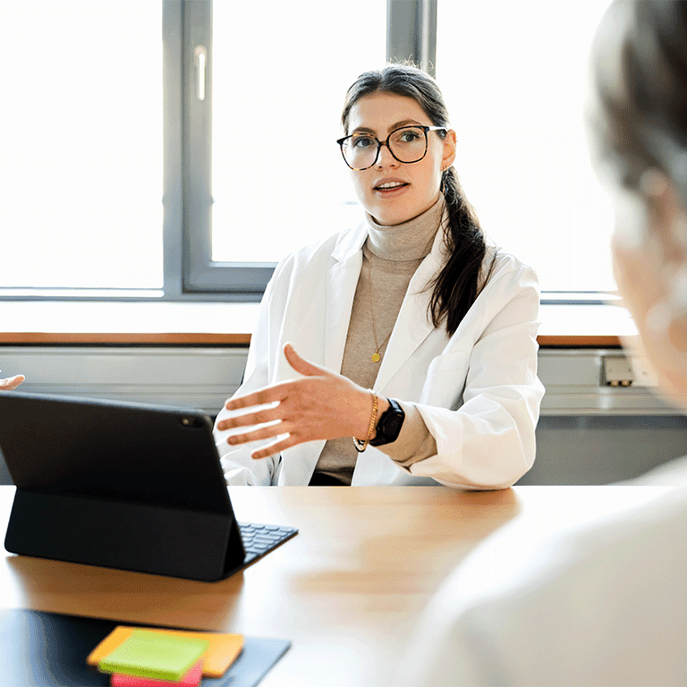 person in a lab coat with a tablet explaining something to someone across the table